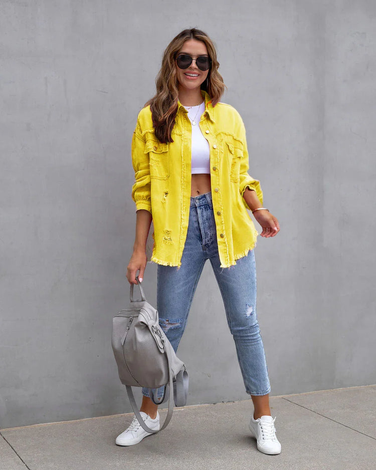 Woman in oversized yellow distressed denim jacket, white crop top, blue ripped skinny jeans, and white sneakers, holding a gray backpack.