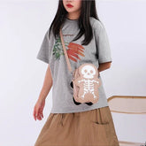 Woman wearing a loose grey graphic t-shirt, tan skirt, and beige skeleton crossbody bag.