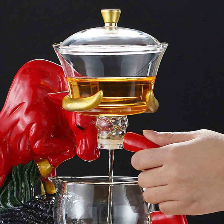 Glass tea pot with gold lid and red bull figurine handle, pouring tea into a clear cup