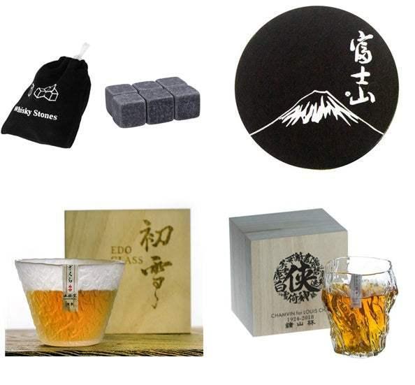 Whiskey stones with pouch, black round coaster with mountain design, and two clear whiskey glasses with amber liquid, each presented in a wooden box.