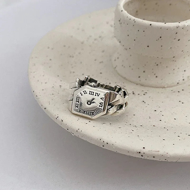 Silver adjustable ring with watch face design and Roman numerals on a ceramic dish