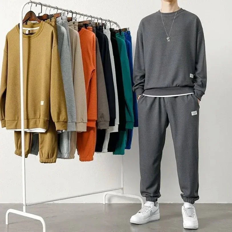Various colored unisex jogger sweat suits on rack, model in loose grey cotton set with white sneakers.