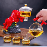 Modern glass teapot set with red bull-shaped base, pouring tea into clear glass cups.