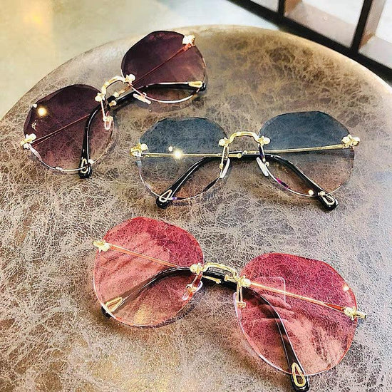 Three pairs of rimless oversized sunglasses with gold stems, gradient pink and gray lenses, laid on a textured brown surface.