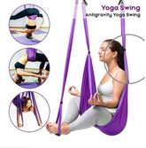 Woman using purple yoga swing hammock for aerial and antigravity poses, wearing grey leggings and a white top.