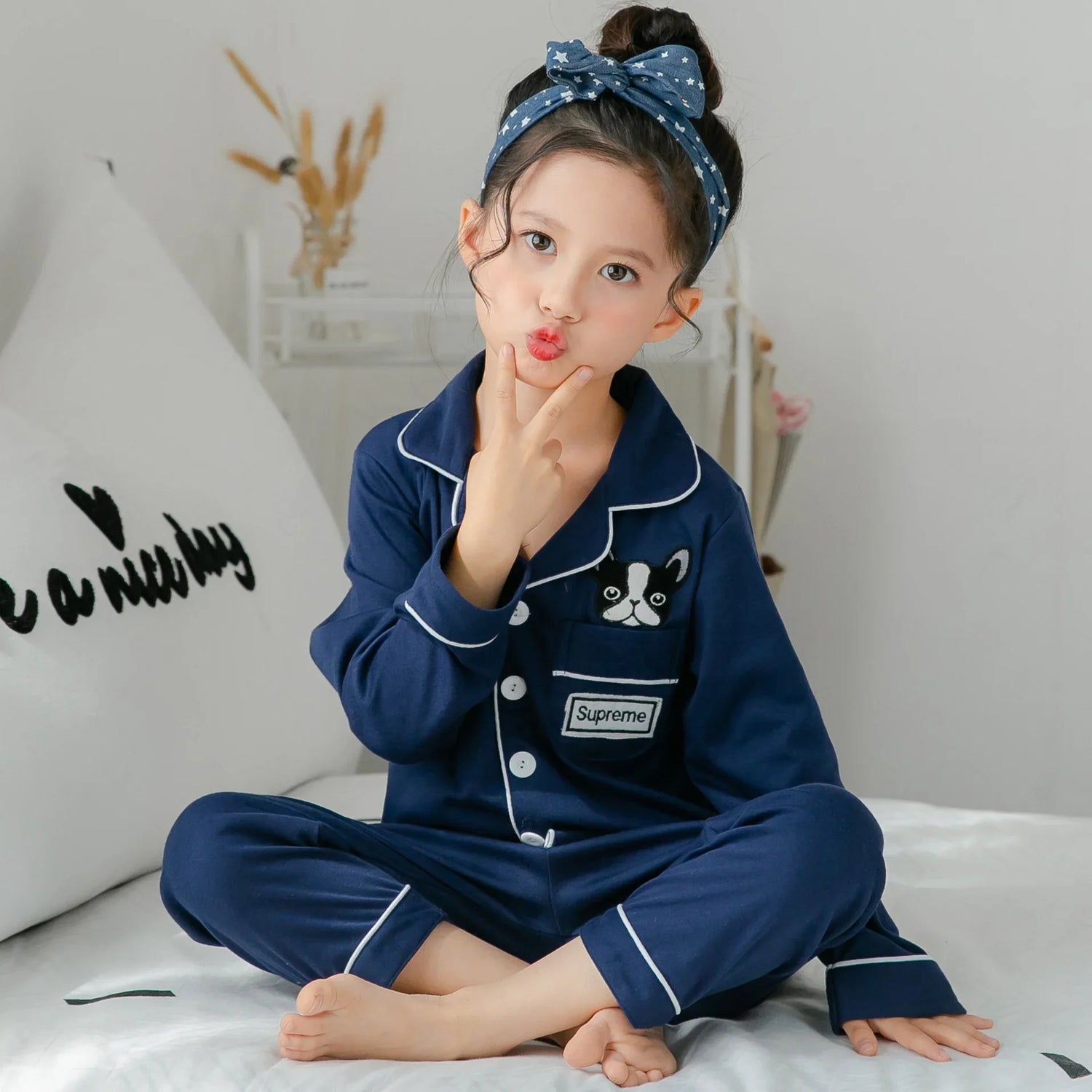 Girl in navy blue cotton pajamas with dog patch, white piping, and star headband, sitting cross-legged.