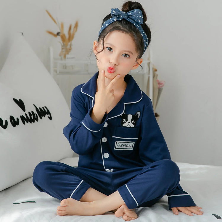 Girl in navy blue cotton pajamas with dog patch, white piping, and star headband, sitting cross-legged.