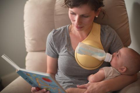 Woman in a casual grey short sleeve shirt using a hands-free baby bottle holder while feeding baby