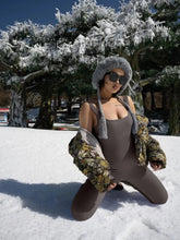 Woman in a fitted brown ribbed jumpsuit, floral puffer jacket, and gray faux fur hat kneeling on snow