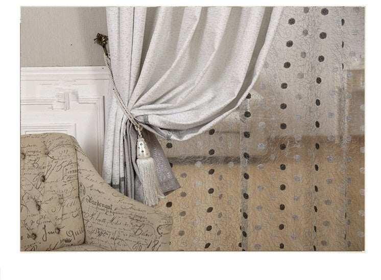 Light grey curtain with sheer polka dot overlay, tied with tassel, next to scripted armchair.