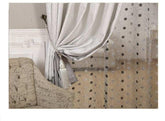 Light grey curtain with sheer polka dot overlay, tied with tassel, next to scripted armchair.