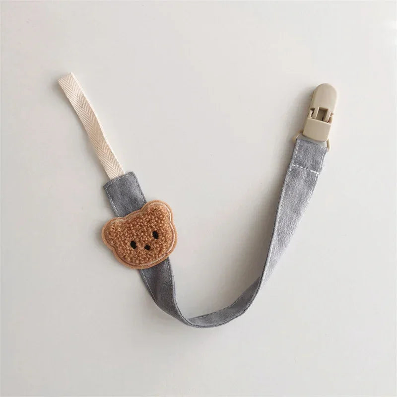 Gray fabric pacifier clip with beige bear patch and cream clip, minimalist style