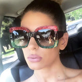Woman wearing oversized square sunglasses with red and green glitter frames, driving in car