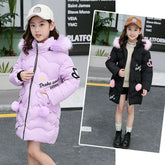 Girl wearing a long padded winter coat with faux fur hood, heart details, and pom-pom accents.