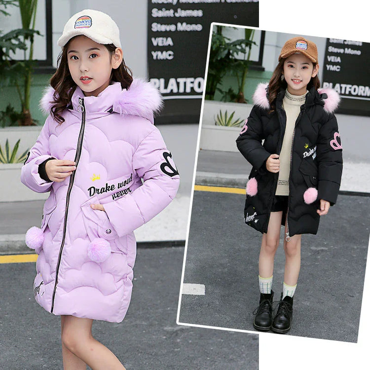 Girl wearing a long padded winter coat with faux fur hood, heart details, and pom-pom accents.