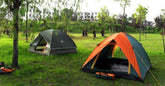Two dome tents, one orange and one green, set up on grassy field with trees, outdoor camping scene.