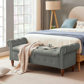 Gray tufted storage bench with rolled arms and wooden legs at the foot of a bed in a bright bedroom