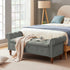 Gray tufted storage bench with rolled arms and wooden legs at the foot of a bed in a bright bedroom