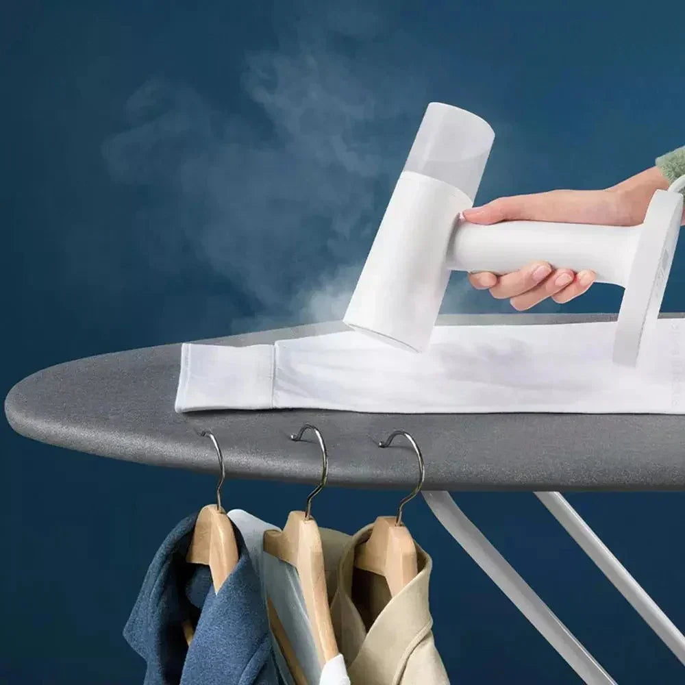 Handheld garment steamer smoothing white long-sleeve shirt on ironing board, casual clothing hanging below.