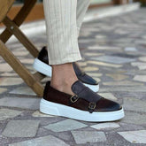 Men’s brown leather slip-on dress shoes with white soles and gold buckles, paired with beige pinstripe pants
