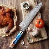 Damascus chef knife with blue resin handle on wooden board, beside tomato, garlic, and roast chicken