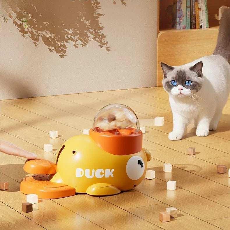 White and gray cat with blue eyes near yellow orange duck-shaped cat treat dispenser on wood floor