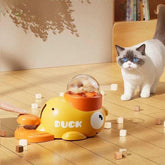 White and gray cat with blue eyes near yellow orange duck-shaped cat treat dispenser on wood floor