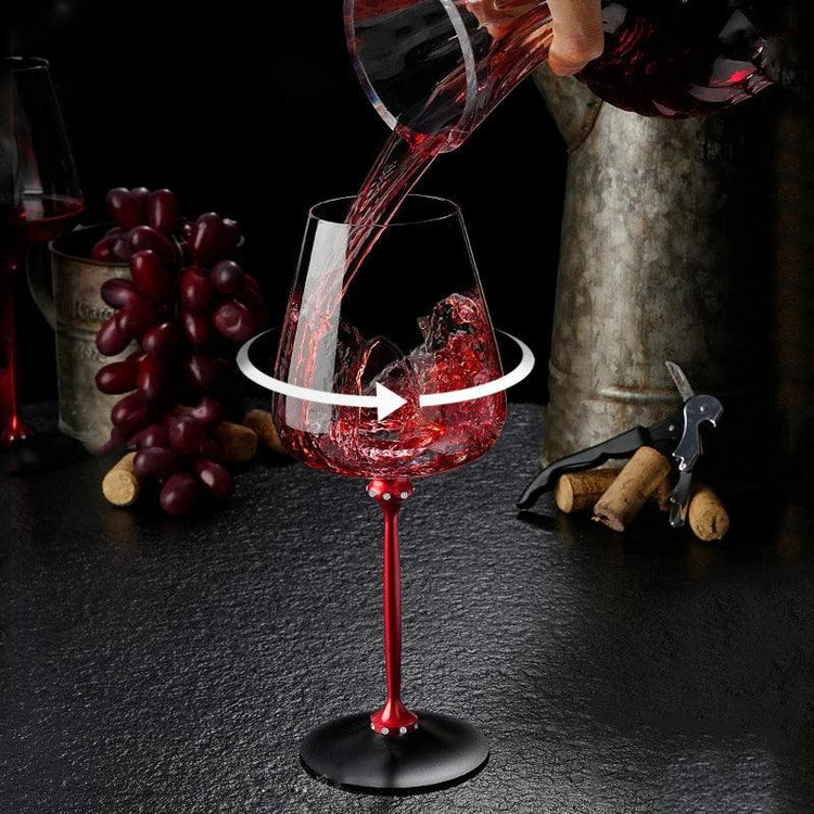 Red wine being poured into a modern glass with a red metallic stem, ice cubes inside, on a dark countertop with grapes and corks nearby.