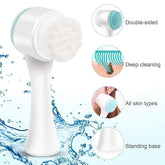 Double-sided white and teal facial cleansing brush with soft bristles, ergonomic handle, and standing base, suitable for all skin types.