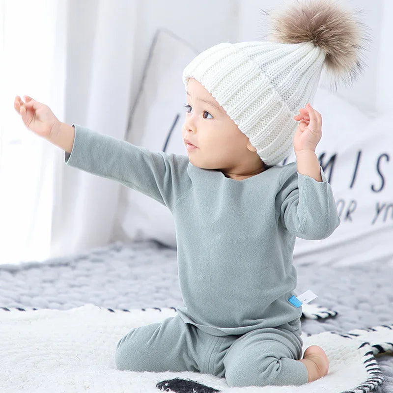 Baby in soft grey fleece loungewear and white knit beanie with faux fur pom sitting indoors