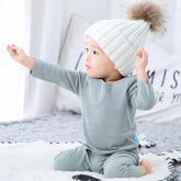 Baby in soft grey fleece loungewear and white knit beanie with faux fur pom sitting indoors