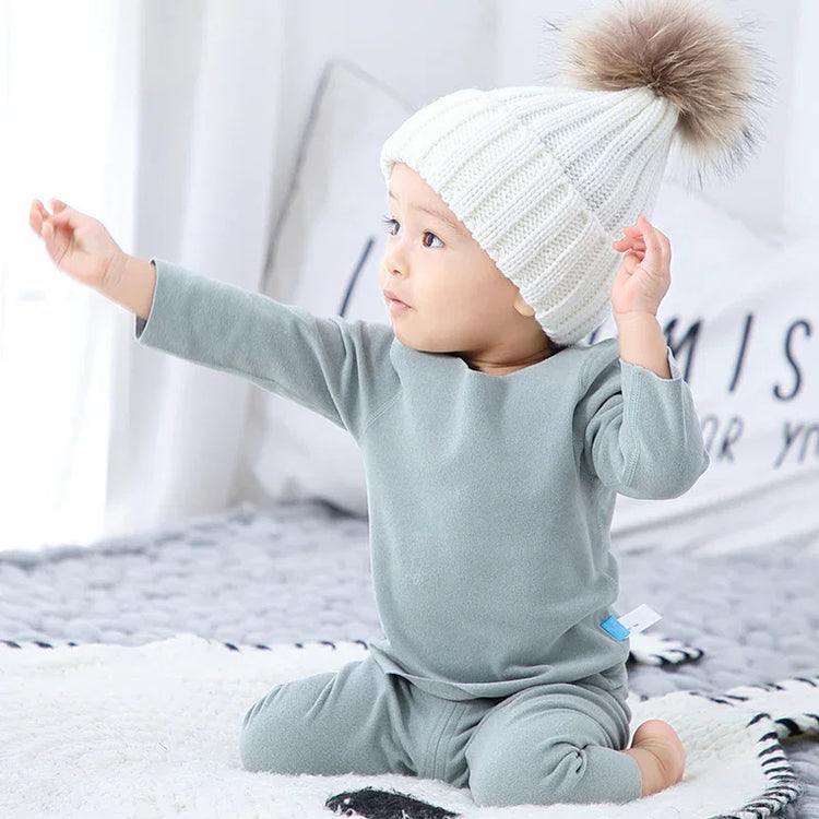 Baby in soft grey fleece loungewear and white knit beanie with faux fur pom sitting indoors