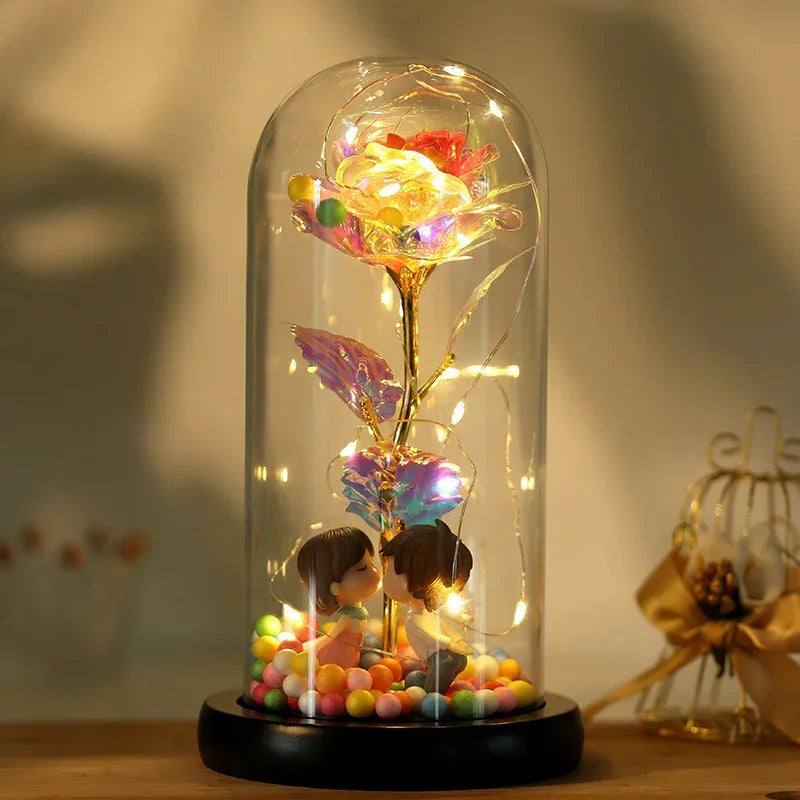Illuminated glass dome holding a gold artificial rose with LED lights and two figurines, surrounded by colorful beads.