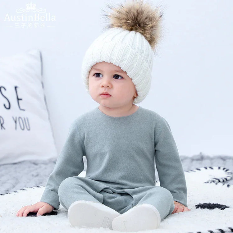 Baby in a light grey long sleeve cotton jumpsuit and chunky white knit beanie with faux fur pom pom, relaxed fit