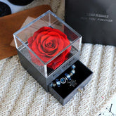 Black gift box with clear lid displaying a red preserved rose and blue charm bracelet in drawer