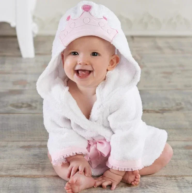 Smiling baby in white hooded bathrobe with pink trim and crown detail, soft plush material, loose fit.