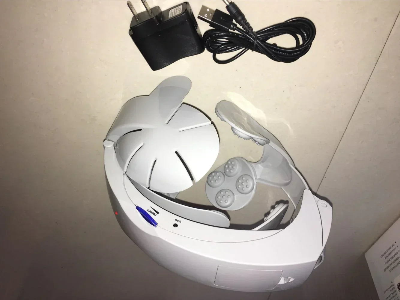 White futuristic plastic head massager with adjustable fit, USB cable and wall charger.