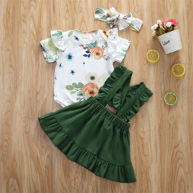 Baby girl floral print romper with ruffled short sleeves, green suspender skirt, and matching headband on wood background.
