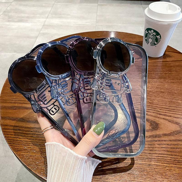 Three transparent astronaut-shaped phone cases with glossy finish in blue, purple, and clear, shown on a wooden table.