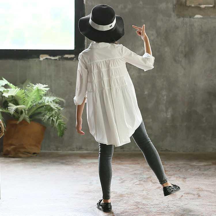 Child wearing long white cotton shirt, grey skinny jeans, black hat, and black flats indoors