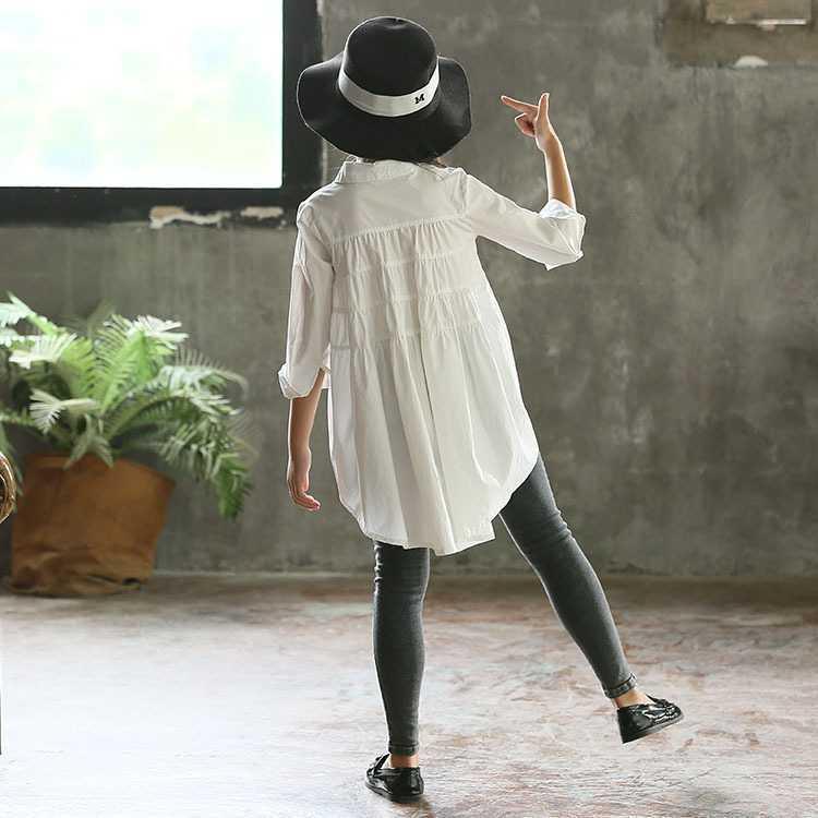Child wearing long white cotton shirt, grey skinny jeans, black hat, and black flats indoors