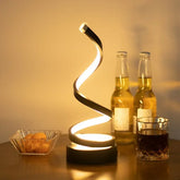 Modern spiral LED table lamp with warm white light on a wooden surface, drinks in background.