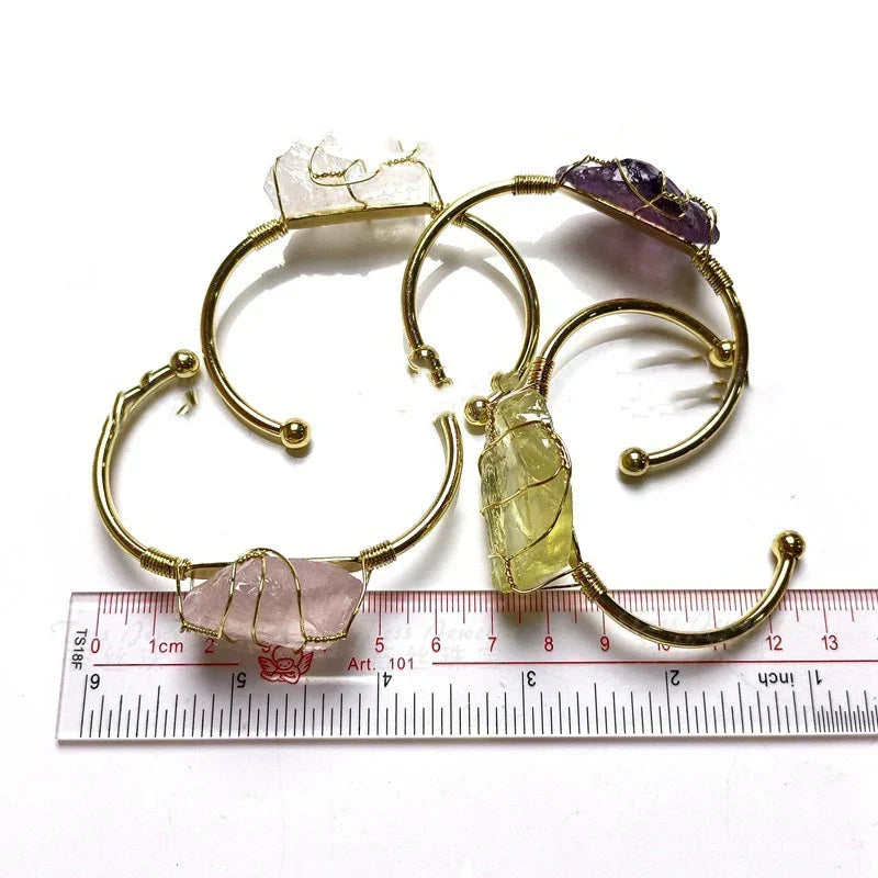Gold open cuff bracelets with raw crystal stones in pink, green, white, and purple, placed beside a ruler