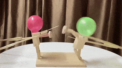 Two wooden stick figure toys with red and green balloon heads, engaging in playful combat on a tabletop