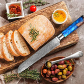 Sliced rustic bread on wooden board with serrated knife, olives, herbs and dipping oil.