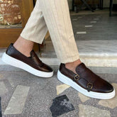 Mens brown leather slip-on sneakers with white soles and double buckle detail, paired with beige pinstripe pants