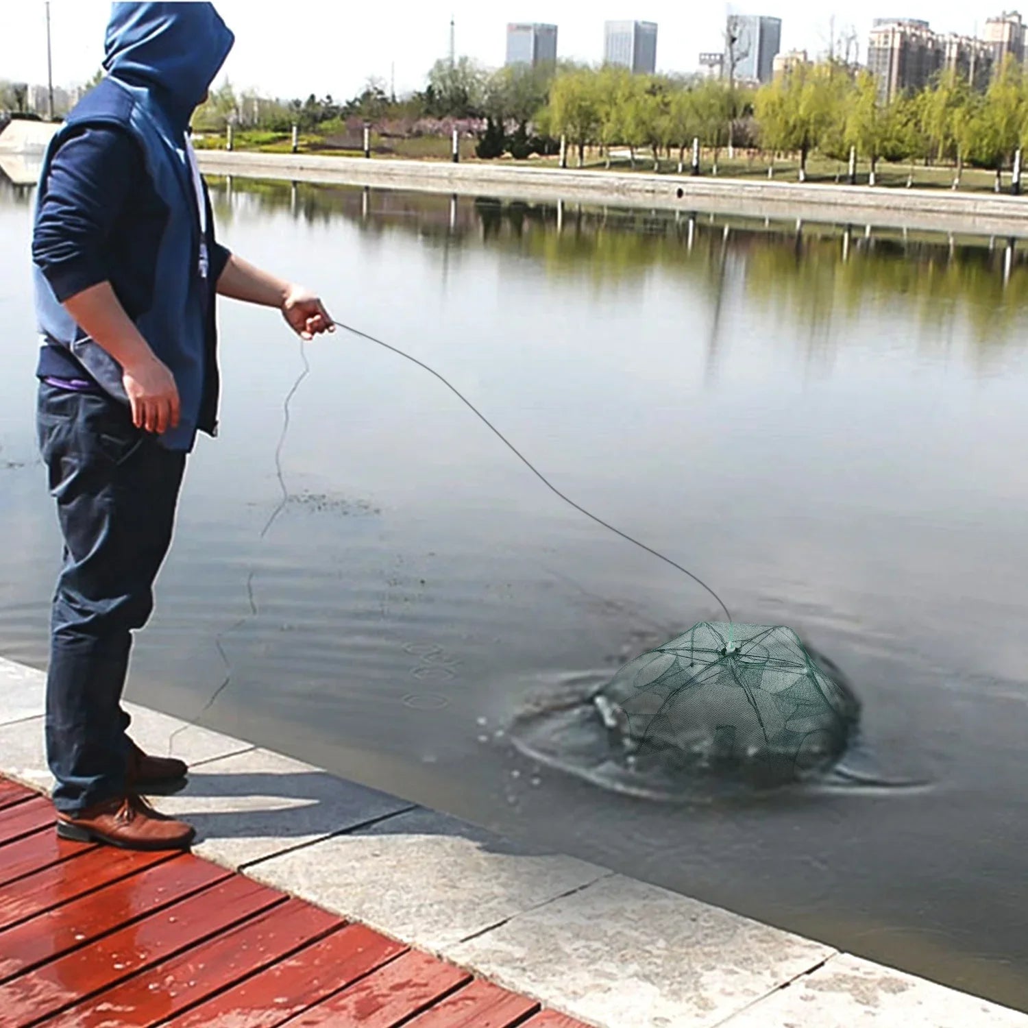 Man in blue hooded jacket and dark pants fishing by a city lake with a collapsible net trap