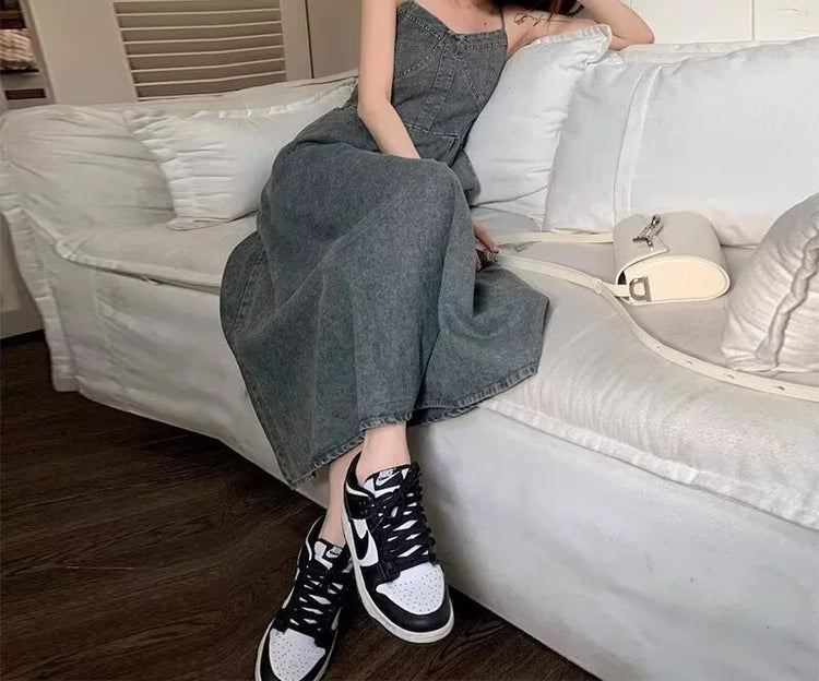 Woman in a sleeveless grey denim maxi dress with relaxed fit, sitting on a sofa, wearing black and white sneakers.