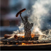 Metal Statue of Liberty grill accessory holding sausage over smoky barbecue, hot dogs grilling