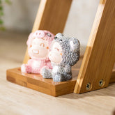 Cute figurines in pink and gray textured animal onesies, sitting on wooden shelf.